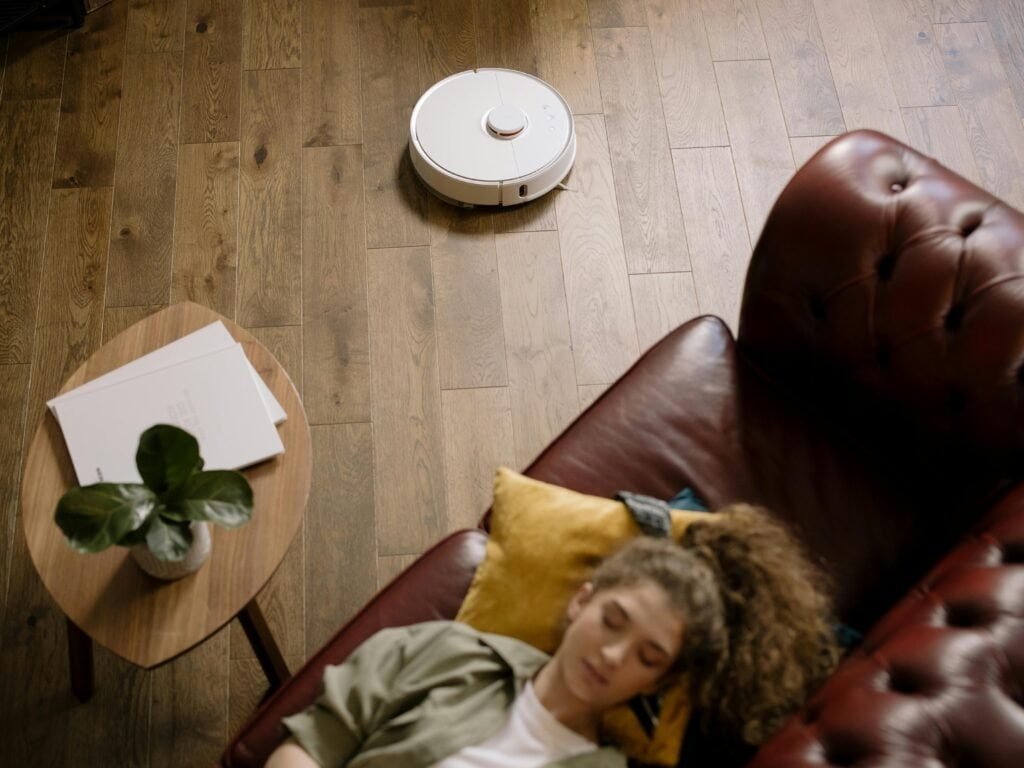 A robot vacuum cleaning a wooden floor while a woman relaxes on a sofa, showing effortless home cleaning.
